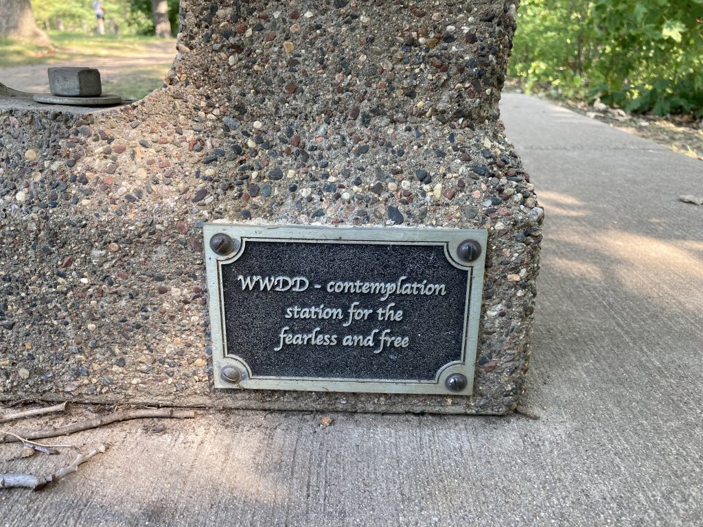 a small plaque at the bottom of a bench. The inscription reads: "WWDD - contemplation station for the fearless and free"