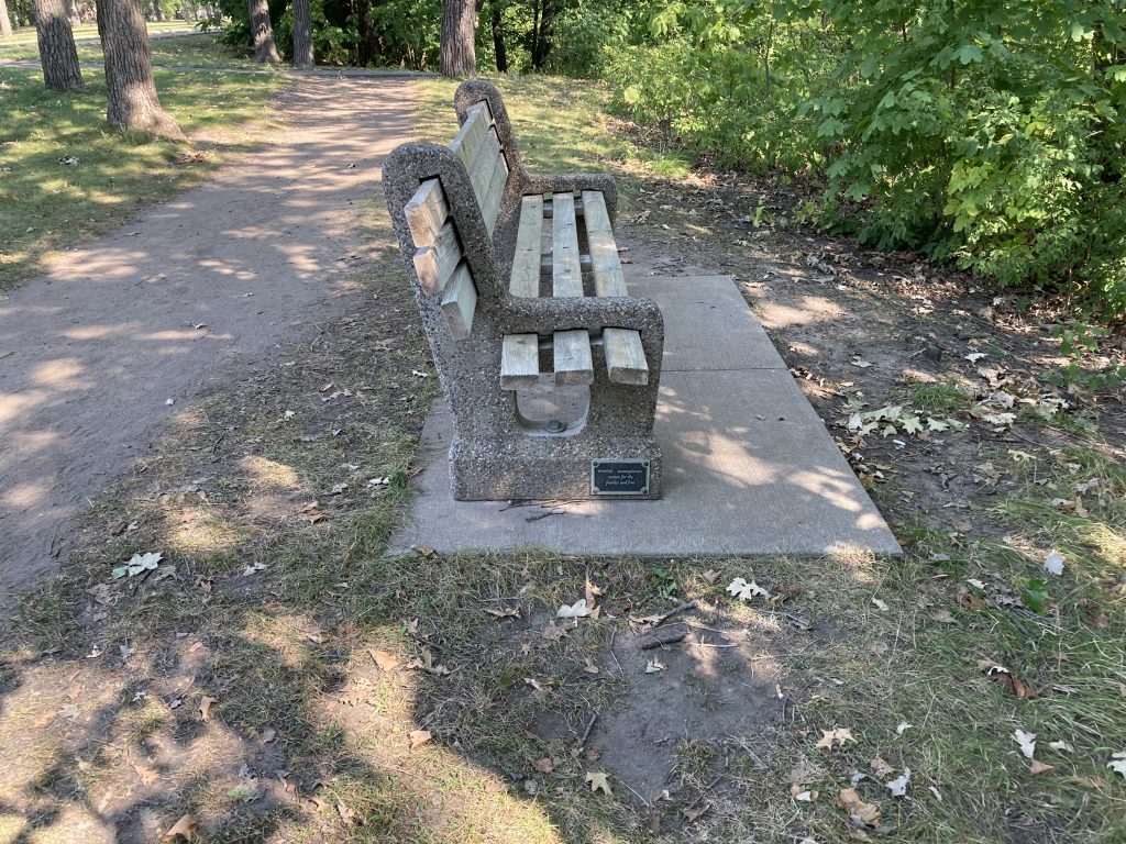 a bench facing trees and the river with a small plaque on it, near the ground