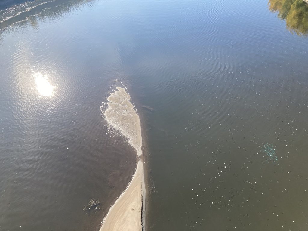 A view from above the river, which is a blue-ish gray, with a white sandbar, strangely shaped, and a shiny circle of sun reflecting on the water. More on the strange shape of the sandbar: in its middle, where it is at its narrowest, the sandbar is covered over with water, making it look like 2 sandbars. Both above and below this spot the sandbar expands.