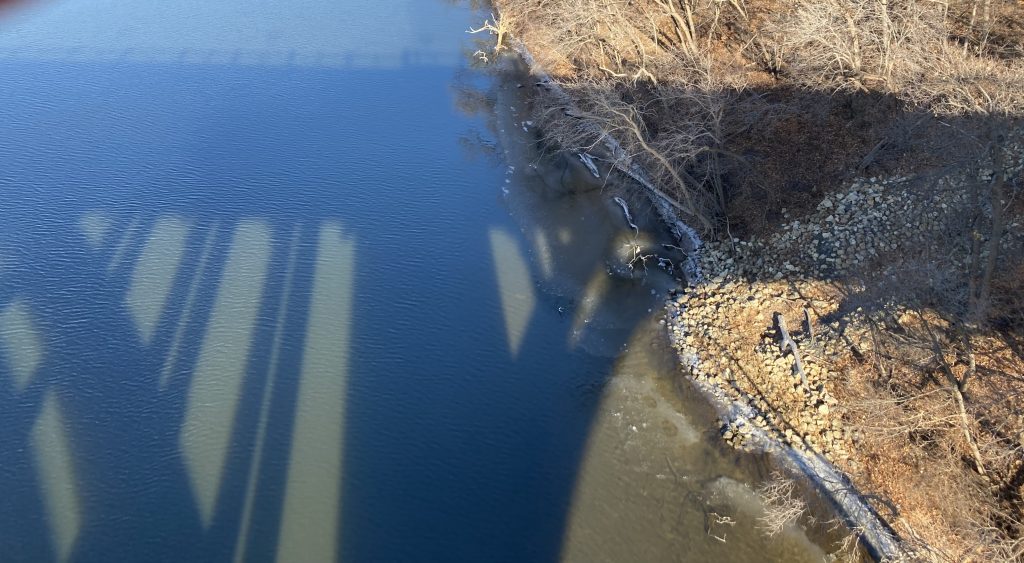 My view from the ford bridge, looking north and down at the Mississippi river. On the right (almost) half of the image is the brownish-greenish shore. On the left, the blue river with dark shadows from the bridge covering it's surface. The shadows are of the columns and are both thick and thin. If I squint hard I can almost see my shadow at the top taking the picture. Is it there, is it just in my imagination?