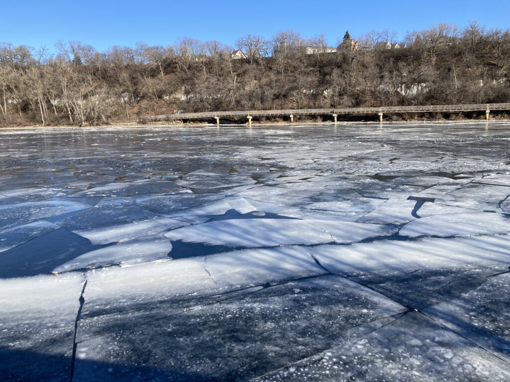 The Mississippi river at the bottom of the franklin hill. White ice, cracks, and shadows on its surface. Beyond it, the east bank with barre branches and blue sky.