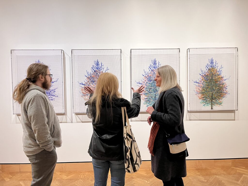 3 people -- a son, a daughter, their mother -- stand in front of a series of trees. The daughter gestures with her hands.
